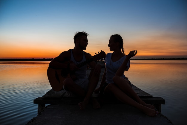 Schattenbilder des jungen schönen Paares, das sich am Sonnenaufgang nahe See erfreut