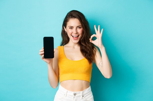 Schau dir das an. Gut aussehende Frau mit frechem Lächeln, Augenzwinkern und Okay-Zeichen mit leerem Smartphone-Bildschirm, App-Empfehlung, auf blauem Hintergrund stehend.