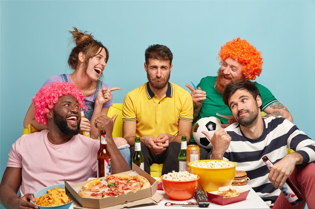 Schau, er ist ein Verlierer. Glückliche Freunde zeigen auf einen unglücklichen Kerl, der andere Teams unterstützt hat, die dieses Match verlieren. Fußballfans haben Spaß, essen leckere Pizza, Popcorn, trinken alkoholische Getränke, schauen sich Großwild an