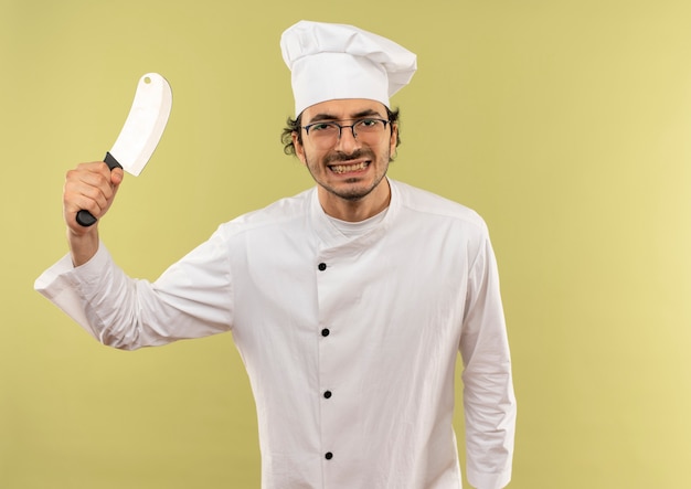 Schauen zufriedener junger männlicher Koch mit Kochuniform und Gläsern, die Hackmesser auf grünem Hintergrund anheben