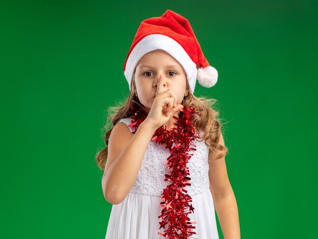 Schauende kleine Mädchen, die Weihnachtsmütze mit Girlande am Hals tragen, die Stille Geste lokalisiert auf grüner Wand zeigt