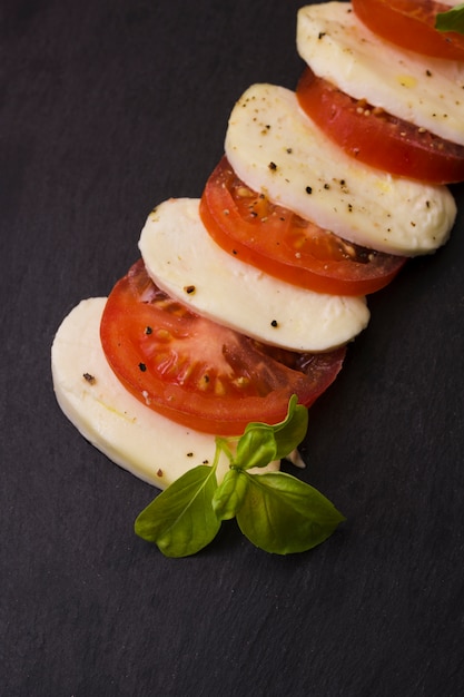 Scheiben von Mozzarellakäse und -tomaten mit Pfeffergewürz und Kräutern auf schwarzem Hintergrund