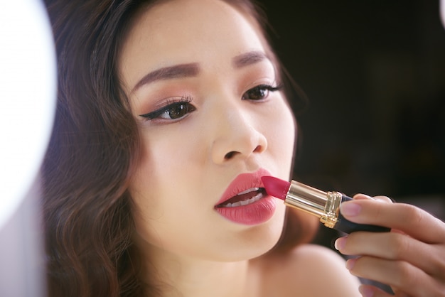 Schicke asiatische Frau, die fertig wird, ihr Lippenmake-up mit rotem Lippenstift zu beenden