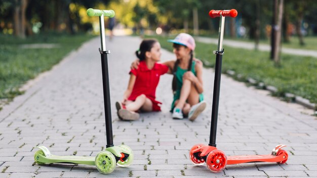 Schieben Sie Roller vor den Mädchen, die auf Gehweg im Park sitzen