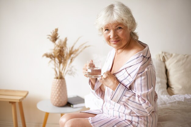 Schläfrige glückliche grauhaarige europäische Rentnerin im stilvollen gestreiften Nachthemd, das im Schlafzimmer auf Bett sitzt und frisches Wasser aus Glas trinkt. Gesunde Gewohnheiten, Alter und Ruhestand