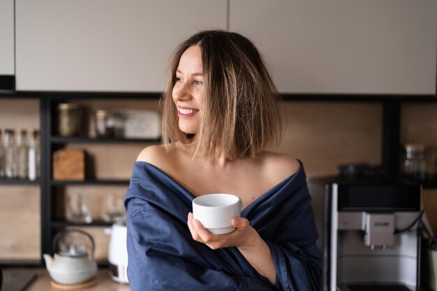 Schläfrige positive Frau, eingewickelt in blaues Bettlaken, die morgens in der Küche Kaffee trinkt