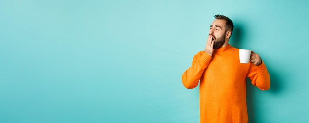 Schläfriger, gutaussehender Mann, der Kaffee trinkt und in orangefarbenem Pullover vor hellblauem Hintergrund gähnt