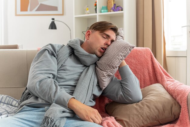 schläfriger junger kranker Mann mit Schal um den Hals, der seinen Kopf auf das Kissen legt, das auf der Couch im Wohnzimmer sitzt