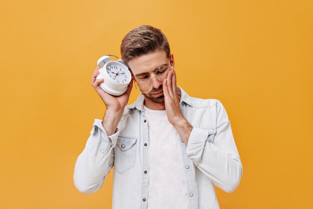 Schläfriger kerl mit braunen haaren in brille, trendiger jacke und hellem t-shirt posiert mit wecker an isolierter wand