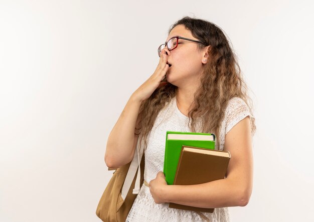 Schläfriges junges hübsches Schulmädchen, das Brille und Rückentasche hält Bücher hält und mit geschlossenen Augen gähnt, die auf weißer Wand isoliert werden