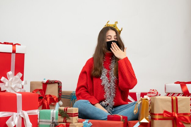 schläfriges junges Mädchen mit rotem Pullover sitzt herum Geschenke mit schwarzer Maske auf Weiß