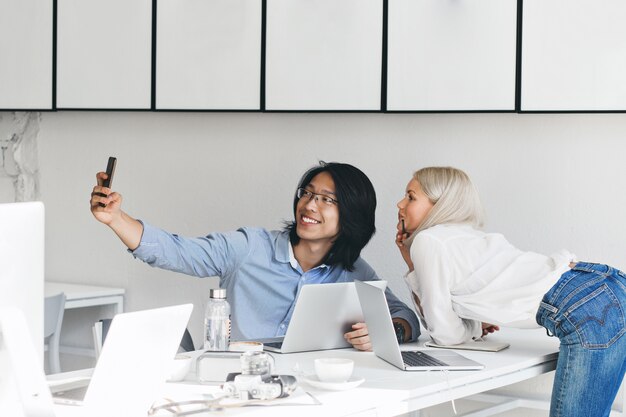 Schlanke blonde Frau im weißen Hemd und in den Jeans, die neben Tisch aufwerfen, während ihr asiatischer Kollege Selfie macht. Innenporträt des chinesischen Arbeiters in den Gläsern, die Spaß mit Sekretärin haben.