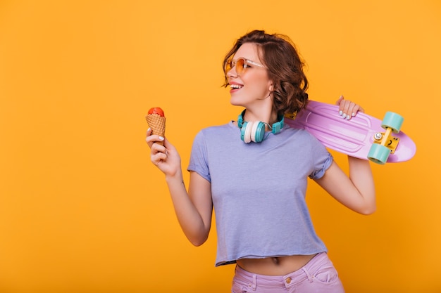 Schlanke europäische Dame mit leckerem Eis lächelnd. Enthusiastisches weibliches Modell mit Skateboard, das Lieblingsdessert genießt.