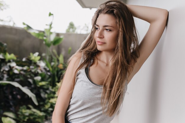 Schlanke europäische Frau mit dunklen Augen, die neben heller Wand aufwerfen. Außenaufnahme des angenehmen Mädchens mit dem braunen Haar, das kühlt.