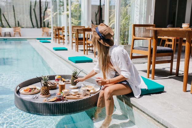 Schlanke Frau im eleganten braunen Hut, der saftige Früchte im Resortcafé isst. Anmutige europäische Frau im weißen Hemd, das mit Cocktail und Essen im Pool entspannt.