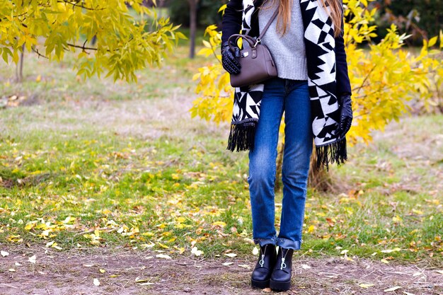 Schlanke Frau trägt blaue Jeans und schwarze Schuhe, die im Herbstwald stehen. Außenporträt des trendigen Mädchens mit dem langen Schal, der mit der kleinen Ledertasche im Oktoberpark aufwirft.