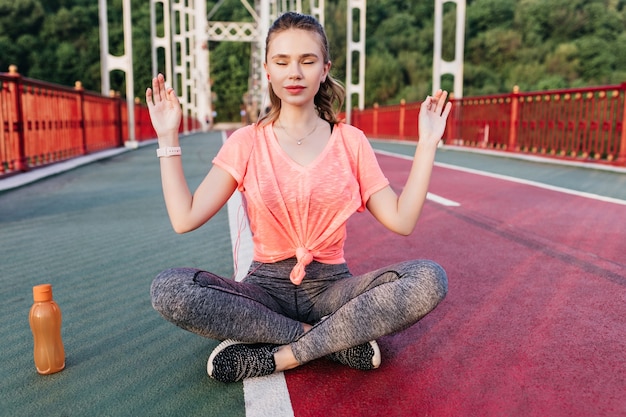 Kostenloses Foto schlankes erstaunliches mädchen, das mit geschlossenen augen an schlackenbahn meditiert. spektakuläre junge dame, die spaß während des outdoor-trainings am sommertag hat.