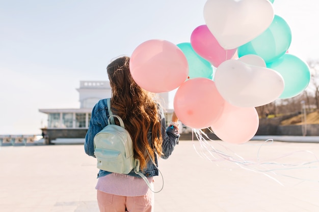 Schlankes Mädchen, das Jeansjacke und stilvolle Hosen trägt, geht nach Party entlang verlassener Böschung. Langhaarige Frau mit niedlichem Rucksack, der Luftballons hält und warmen Wind weht.