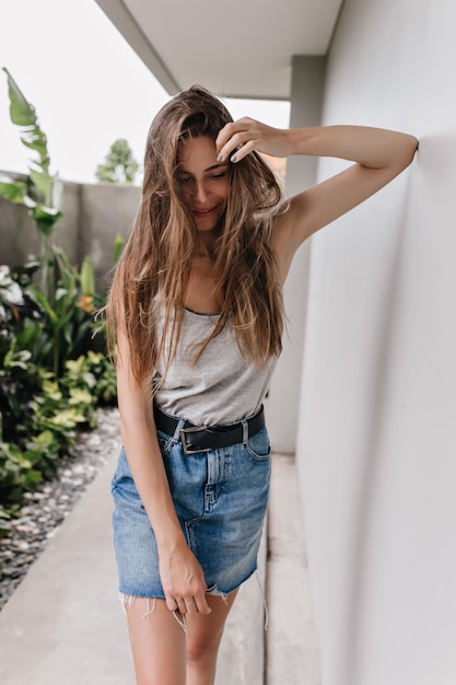 Schlankes Mädchen trägt eleganten Rock, der mit schüchternem Lächeln aufwirft. Außenporträt der siegreichen Dame mit dem langen Haar, das nahe weißer Wand steht.