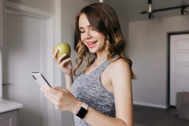 Schlankes reizendes Mädchen mit Smartphone am Morgen. Innenporträt der atemberaubenden dunkelhaarigen Frau, die grünen Apfel isst und lächelt.