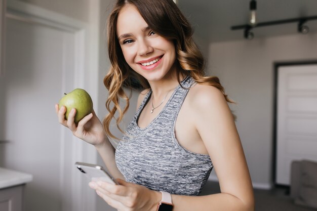 Schlankes wunderschönes Mädchen mit welligem Haar, das mit Telefon und Obst aufwirft. Innenporträt des romantischen weiblichen Modells, das grünen Apfel am Morgen isst.