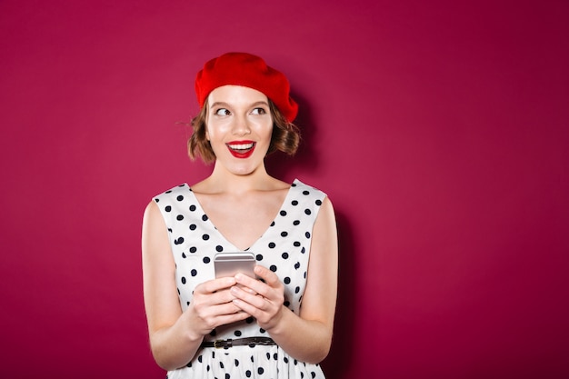 Schlaue lächelnde Ingwerfrau im Kleid, das Smartphone hält und weg über Rosa schaut