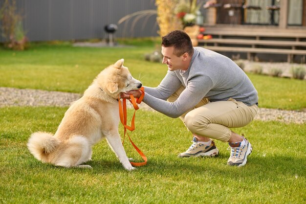 Schlauer Hund. Seitlich zur Kamera eines fröhlichen jungen erwachsenen Mannes, der am Herbsttag in der Nähe des Landhauses hockt und einen freundlichen und klugen Hund mit Leine berührt