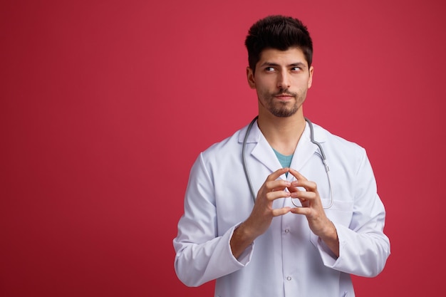 Schlauer junger männlicher Arzt mit medizinischer Uniform und Stethoskop um den Hals, der die Hände zusammenhält und die Seite isoliert auf rotem Hintergrund mit Kopierraum betrachtet
