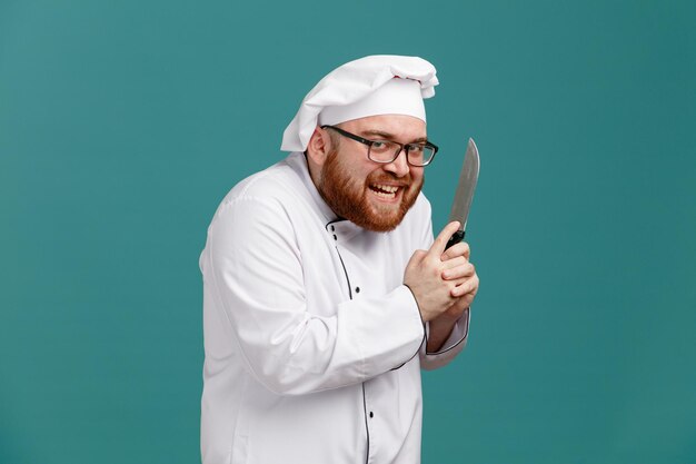 Schlauer junger männlicher Koch mit Brille, Uniform und Mütze, der in der Profilansicht steht und ein Messer mit beiden Händen hält, die in die Kamera schauen und eine Pistolengeste machen, die etwas Böses isoliert auf blauem Hintergrund plant