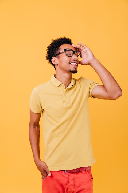 Schlauer Kerl im lässigen Outfit, das mit geschlossenen Augen aufwirft. Innenfoto des angenehmen afrikanischen jungen Mannes in den Gläsern.