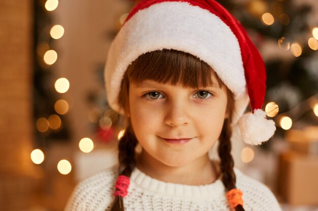 Schließen Sie das Porträt eines süßen, charmanten weiblichen Kindes mit weißem Pullover und Weihnachtsmann-Hut, das die Kamera mit positivem Ausdruck betrachtet und in guter festlicher Stimmung ist.