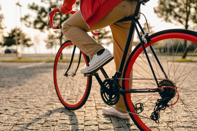 Schließen Sie die Beine in Turnschuhen und Händen am Lenkrad des bärtigen Mannes im Hipster-Stil im roten Kapuzenpulli und in der beige Hose, die allein mit Rucksack auf Fahrrad reisenden Rucksackreisenden des gesunden aktiven Lebensstils reiten