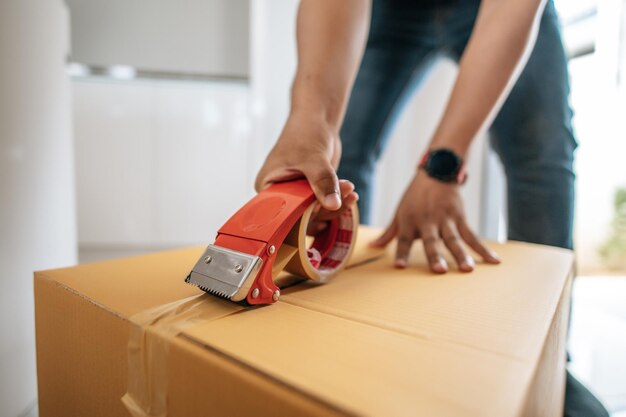 Schließen Sie die Hand des Menschen mit Klebeband, um den Karton zu schließen, der eine große Kiste zum Verpacken des neuen Hauskonzepts versiegelt