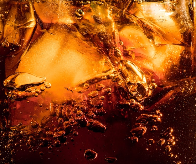 Schließen Sie herauf Ansicht der Eiswürfel im dunklen Cola-Hintergrund. Textur des kühlenden süßen Sommergetränks mit Schaum und Makroblasen an der Glaswand. Zischen oder Schweben bis zur Oberfläche.