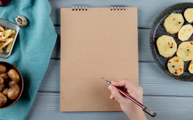 Schließen Sie herauf Ansicht der Frauenhand, die Stift und Notizblock mit gebackenen und geschnittenen Kartoffeln herum auf hölzernem Hintergrund mit Kopienraum hält