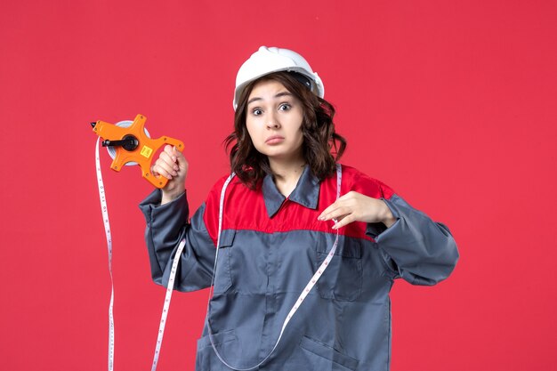 Schließen Sie herauf Ansicht des schockierten emotionalen weiblichen Architekten in der Uniform, die Schutzhelm mit Maßband auf roter Wand trägt