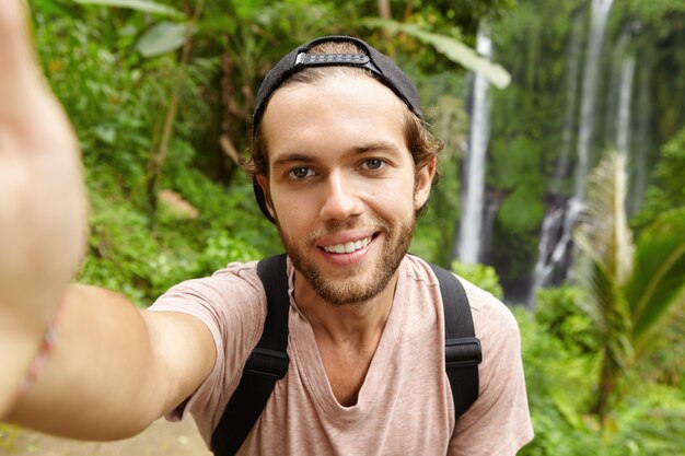 Schließen Sie herauf Ansicht des schönen kaukasischen Wanderers, der Hysterese trägt, die mit glücklichem Lächeln schaut, während Selfie mit erstaunlicher Landschaft mit Wasserfall nimmt