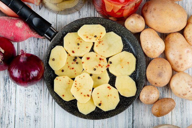 Schließen Sie herauf Ansicht von Kartoffelscheiben mit schwarzem Pfeffer in Pfanne und Zwiebelrettichkartoffeln herum auf hölzernem Hintergrund