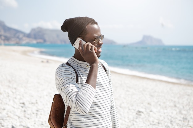Schließen Sie herauf Außenporträt des schwarzen Rucksacktouristen im Hut, der am Strand steht und per Telefon spricht