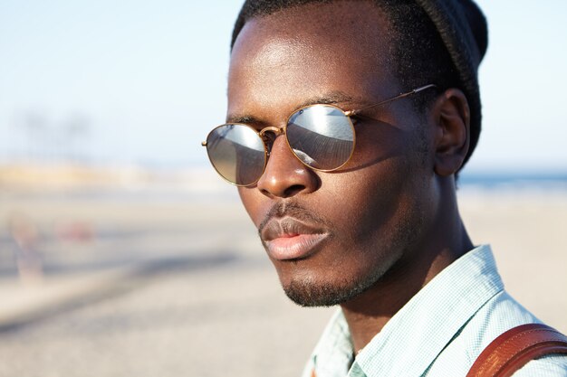 Schließen Sie herauf Außenporträt des selbstbewussten attraktiven jungen afroamerikanischen Mannes in der Sonnenbrille der verspiegelten Linse, die Morgen am Meer verbringt, sich entscheidet, zögernd über die Wahl des harten Lebens