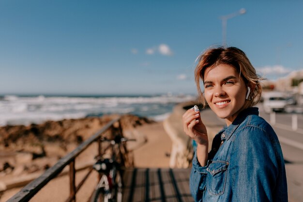 Schließen Sie herauf außerhalb des Porträts der reizenden lächelnden Frau, die Jeanshemd hält, das Airpods hält und auf den Ozean im sonnigen Tag schaut