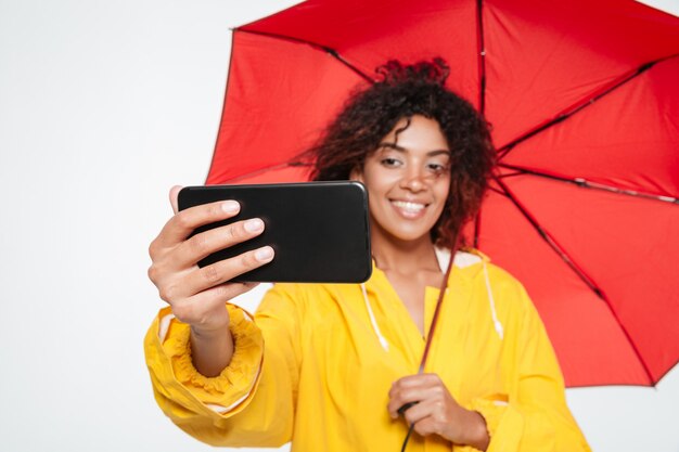 Schließen Sie herauf Bild der lächelnden afrikanischen Frau im Regenmantel, der sich unter Regenschirm versteckt und Selfie auf ihrem Smartphone über weißem Hintergrund macht