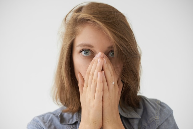 Schließen Sie herauf Bild der verängstigten jungen Frau mit der losen Frisur, die Gesicht mit beiden Händen bedeckt und mit ängstlichem Blick in die Kamera starrt, gestresst oder mit etwas erschrocken ist. Menschliche Gefühle