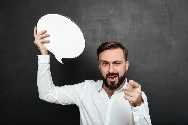 Schließen Sie herauf das Foto des überzeugten strengen Mannes, der leere Spracheblase hält und Zeigefinger auf Kamera über dunkelgrauem Wandkopienraum zeigt