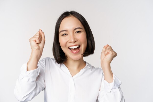 Schließen sie herauf das gesichtsporträt der tanzenden asiatischen frau, die triumphierend lächelt und mit dem glücklichen gefühl feiert, das über weißem hintergrund steht