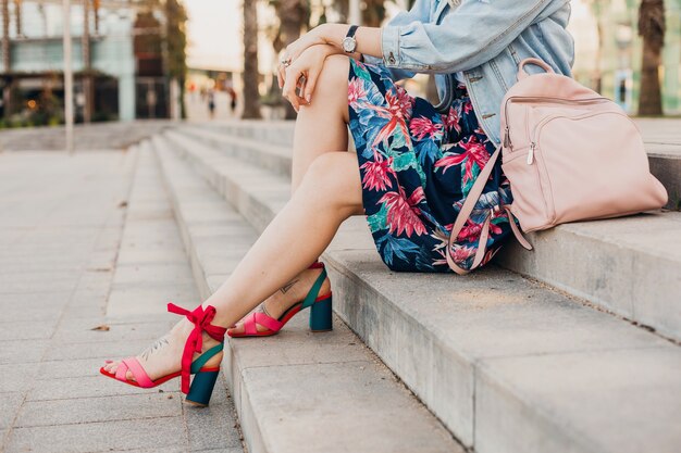 Schließen Sie herauf Details der Beine in den rosa Sandalen der Frau, die auf Treppen in der Stadtstraße im stilvollen bedruckten Rock mit Lederrucksack, Sommerarttrend sitzt