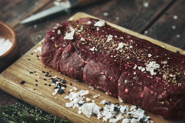 Schließen Sie herauf fokussiertes Stück Fleisch gesalzen, das auf Holzbrett auf Vintage-Tisch zwischen Gewürzen gepfeffert wird