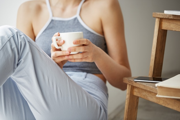 Schließen sie herauf foto des alten büchertelefons, das auf stuhl liegt, frau, die auf boden sitzt und tasse kaffee über weißer wand hält.