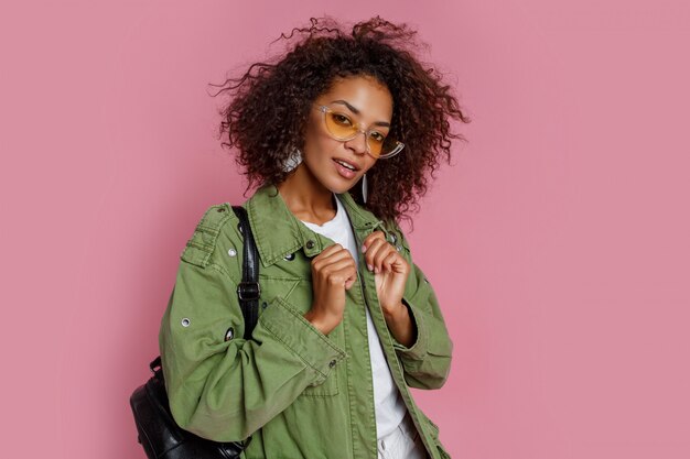 Schließen Sie herauf Foto des erstaunlichen lockigen afrikanischen Mädchens auf rosa Hintergrund. Trägt eine grüne, trendige Jacke.