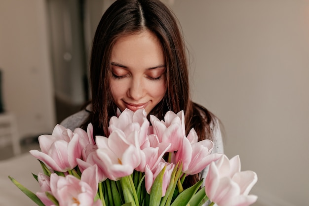 Schließen Sie herauf Innenporträt der glücklichen Frau mit dunklem Haar, das mit geschlossenen Augen und glücklichem Lächeln, das Blumen hält, aufwirft.